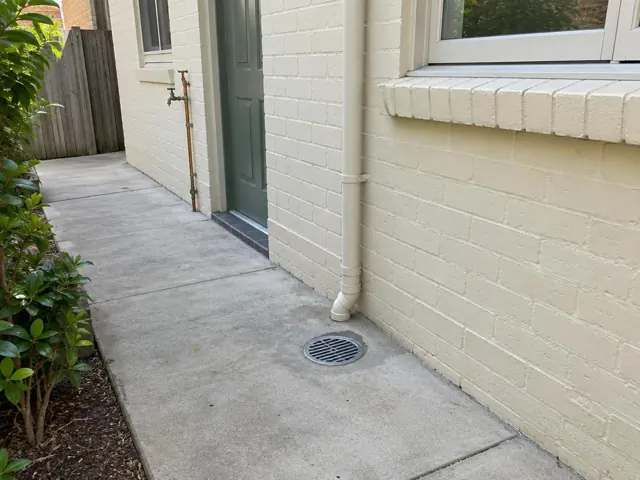 Outdoor drain gully grate beside a cream brick wall and concrete path in Manly