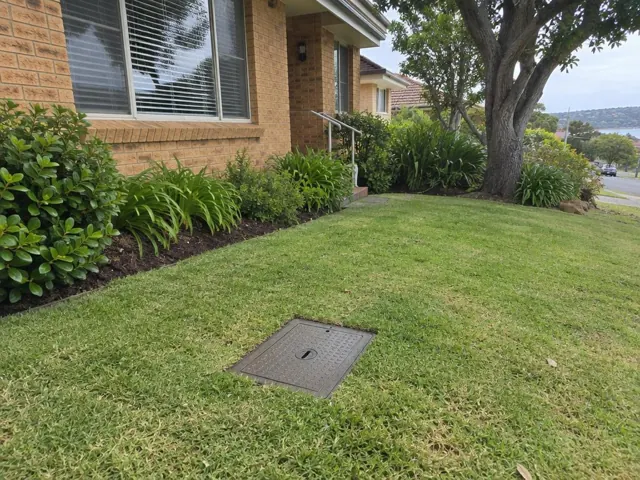 Flush square sewer access cover in a tidy front lawn beside a brick house in Curl Curl